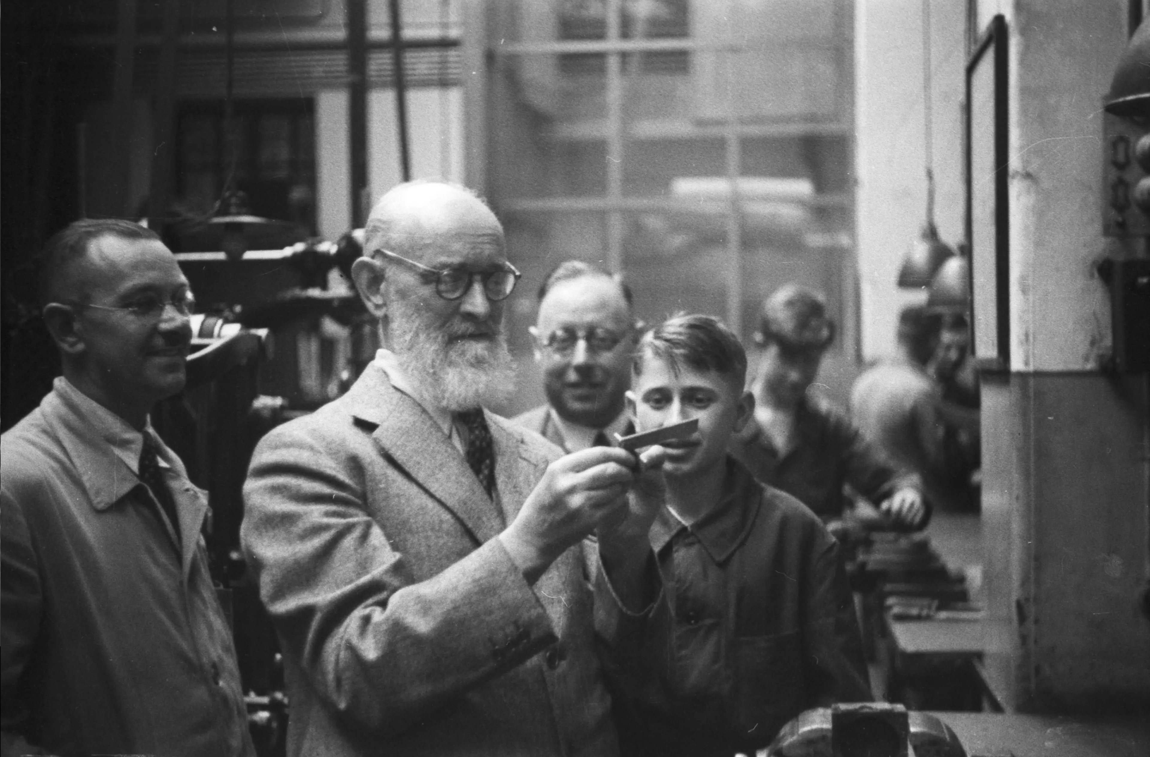 Historical black and white photograph of Robert Bosch with a beard and glasses in a workshop. He is surrounded by employees and is inspecting a small workpiece.