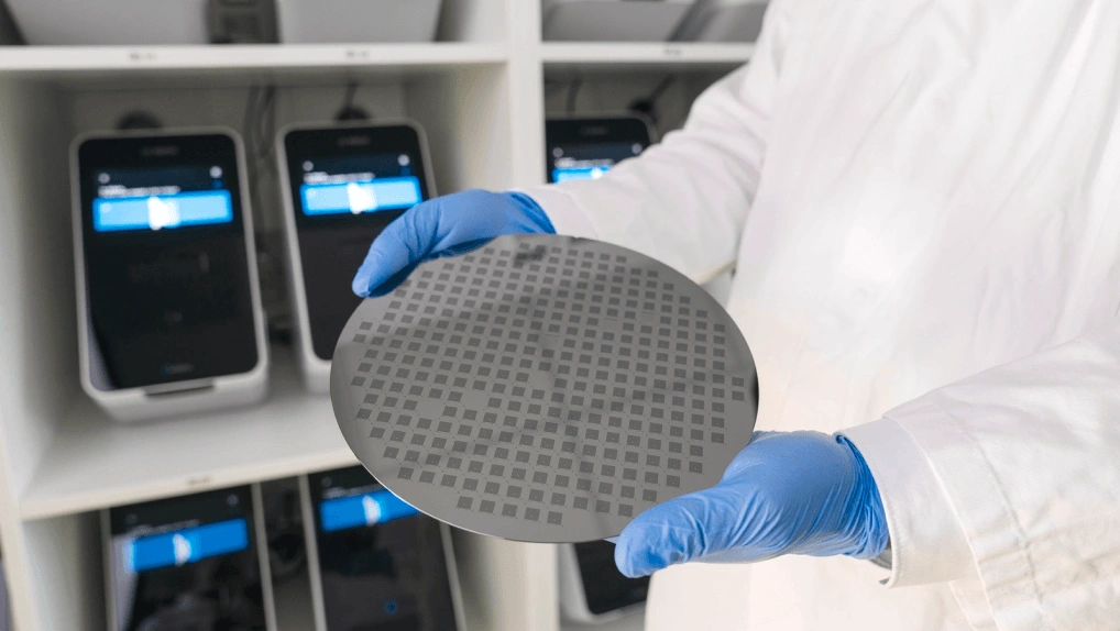 A person in a white lab coat and blue disposable gloves holds a circular silicon wafer showing numerous microchips. In the background are medical analysis devices.