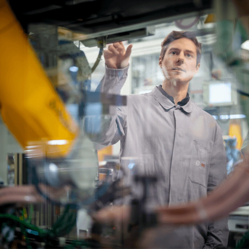 A Bosch employee in a grey work coat stands in a production facility looking at a machine. The photo is taken through a glass panel, which reflects a yellow robotic arm.
