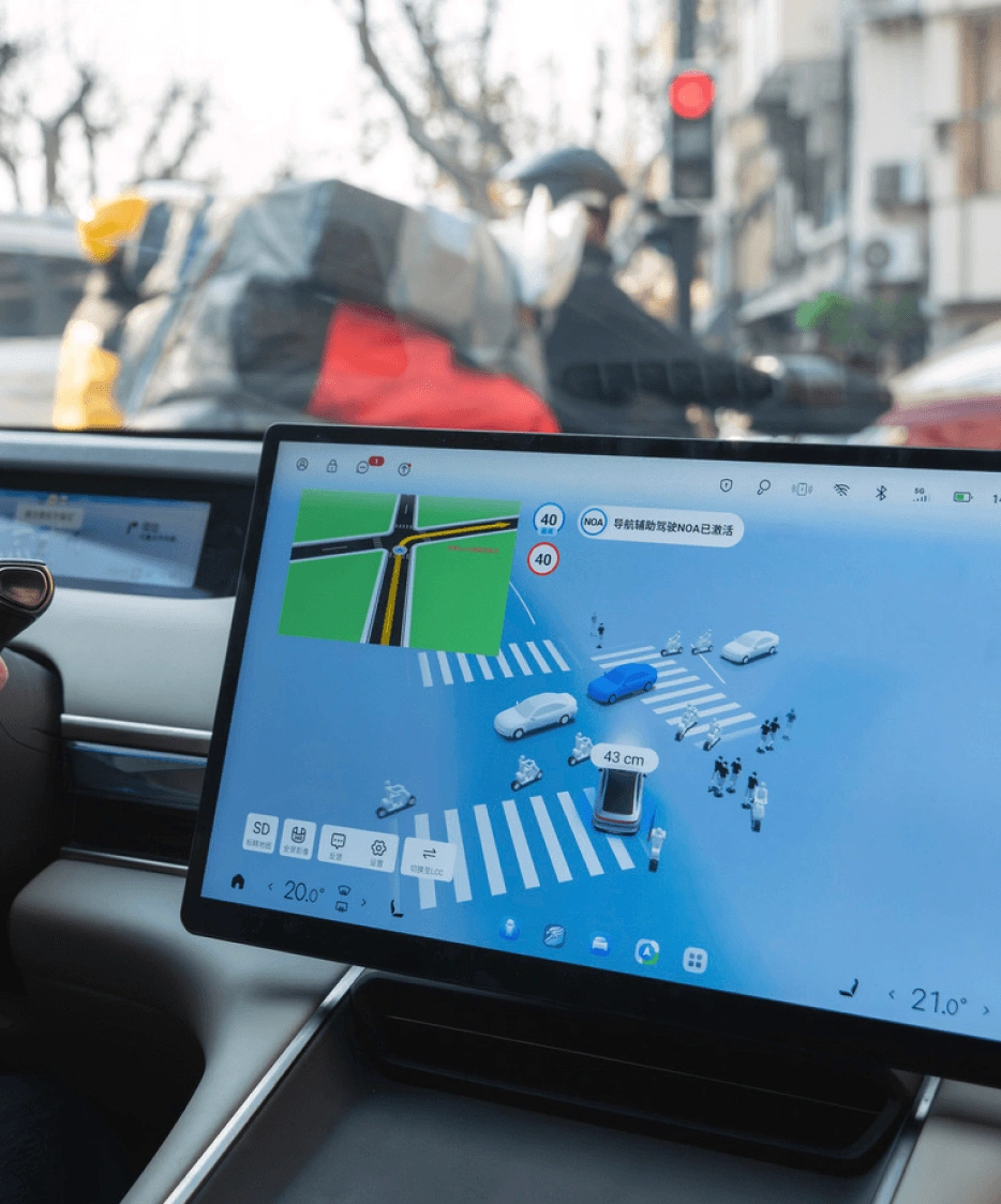 Close-up of a car's dashboard. The driver's hands are on the steering wheel. A large central touchscreen displays the ADAS system, a 3D visualization of the car's surroundings, including other road users and lane markings.