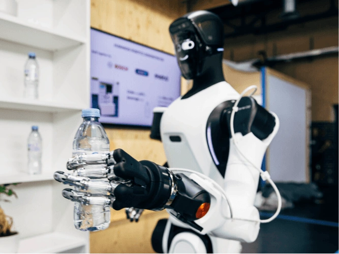 A humanoid robot with a white and black body holds a plastic water bottle with its metallic robotic hand. In the background are white shelves.